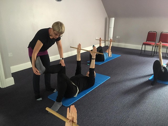 a group of pilates students on their backs, raising poles about their heads while also doing leg exercises